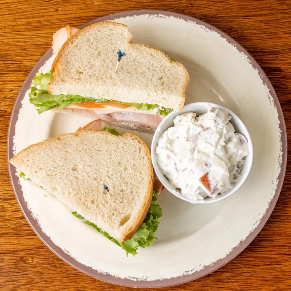 Turkey sandwich with lettuce and tomato, served with a side of potato salad on a plate.