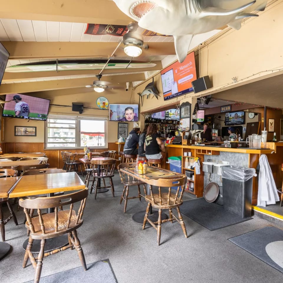 Casual restaurant interior with wooden tables, chairs, a bar, and TVs, featuring a large shark decoration.