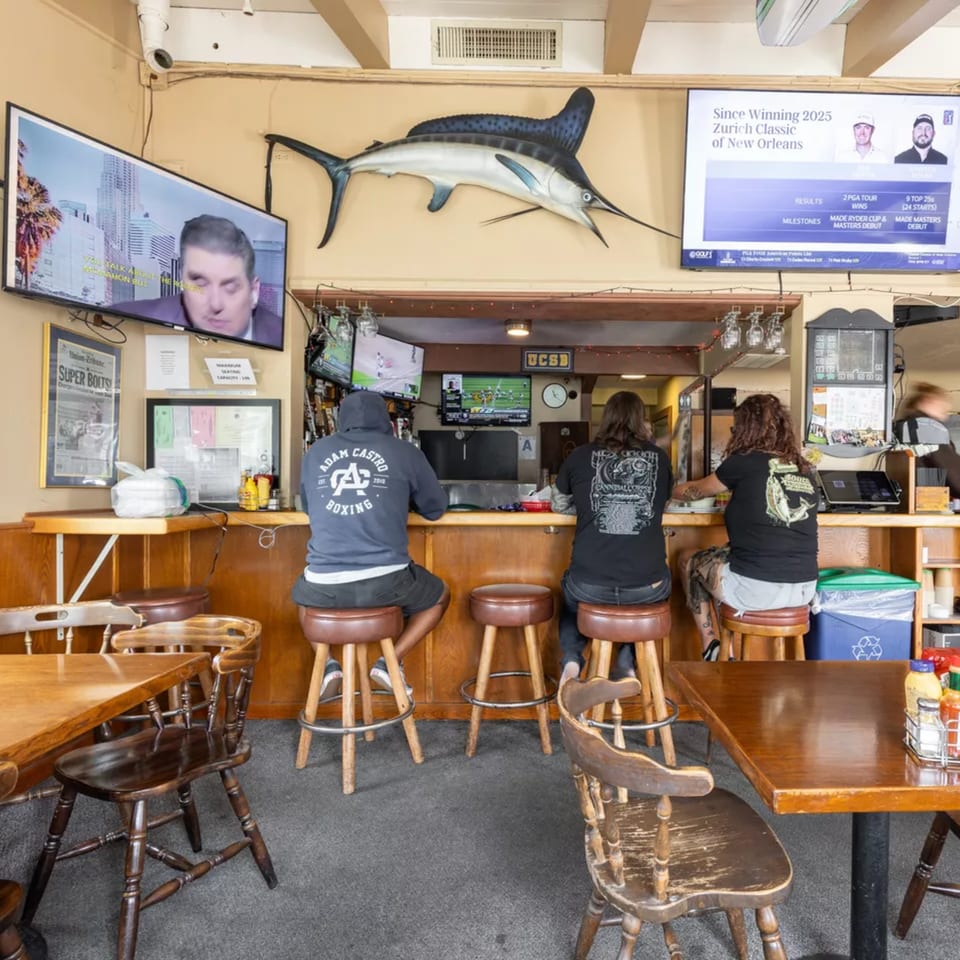 Three people sit at a bar with TVs showing sports, a marlin decoration, and wooden tables.