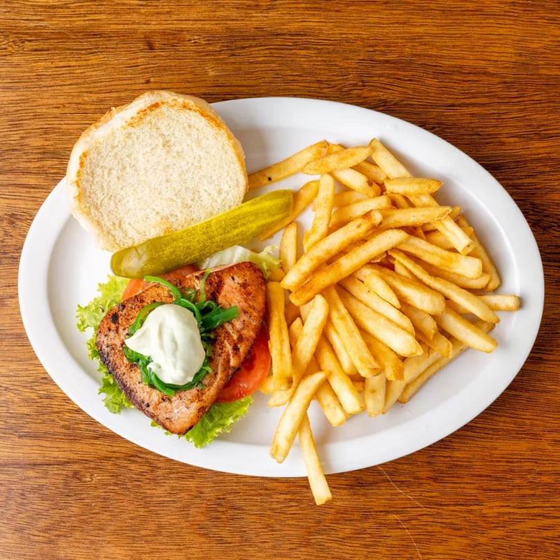Grilled salmon sandwich with lettuce, tomato, and sauce, served with french fries and a pickle.