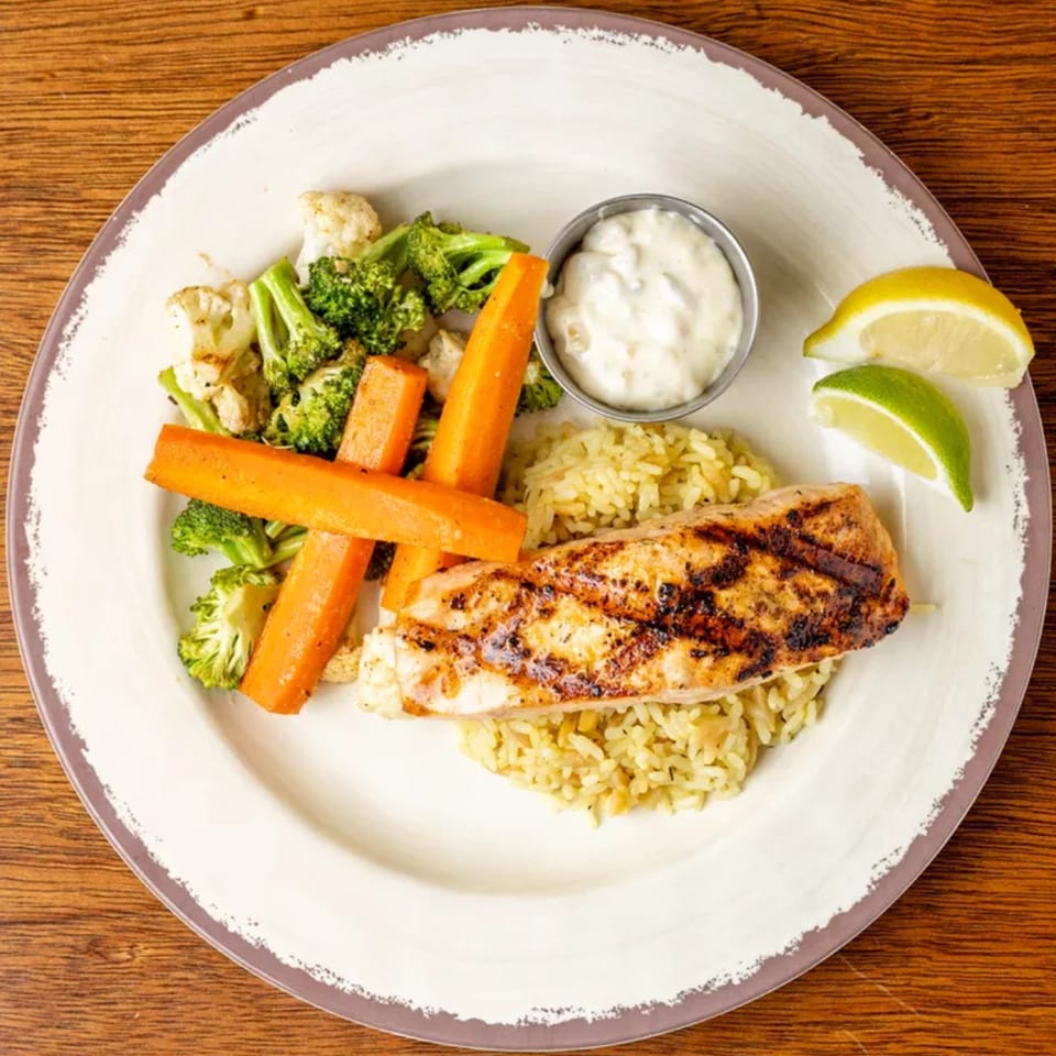Grilled salmon with rice, roasted carrots, broccoli, cauliflower, a side of sauce, and citrus wedges.