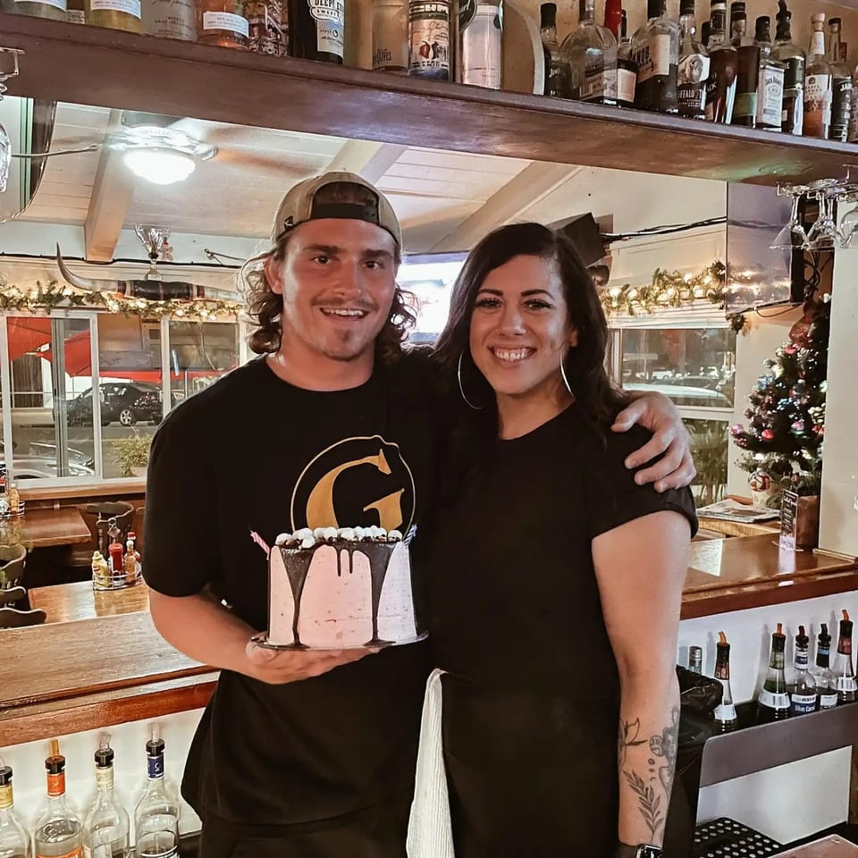 Two smiling people, a man and a woman, stand behind a bar holding a pink cake.