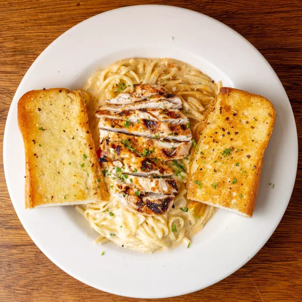 Grilled chicken Alfredo pasta with two slices of garlic bread on a white plate.