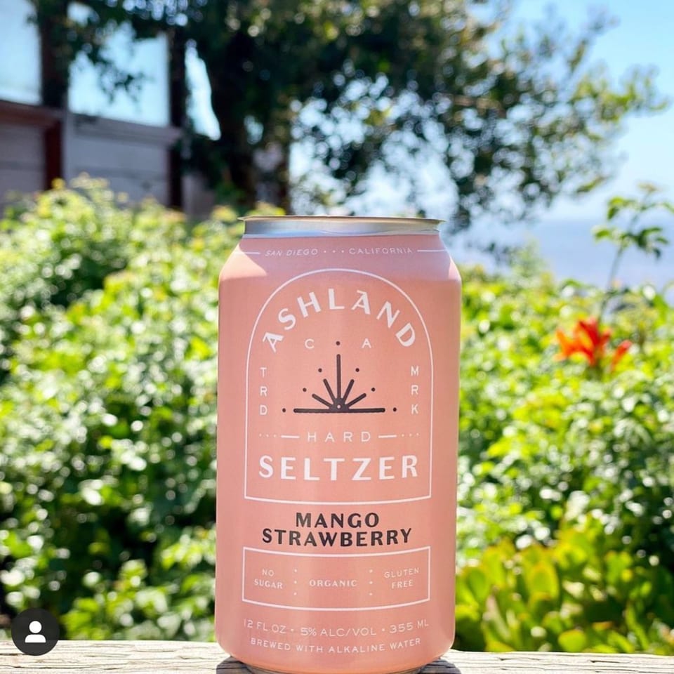 Ashland Hard Seltzer Mango Strawberry can on a wooden surface with a blurred green background.