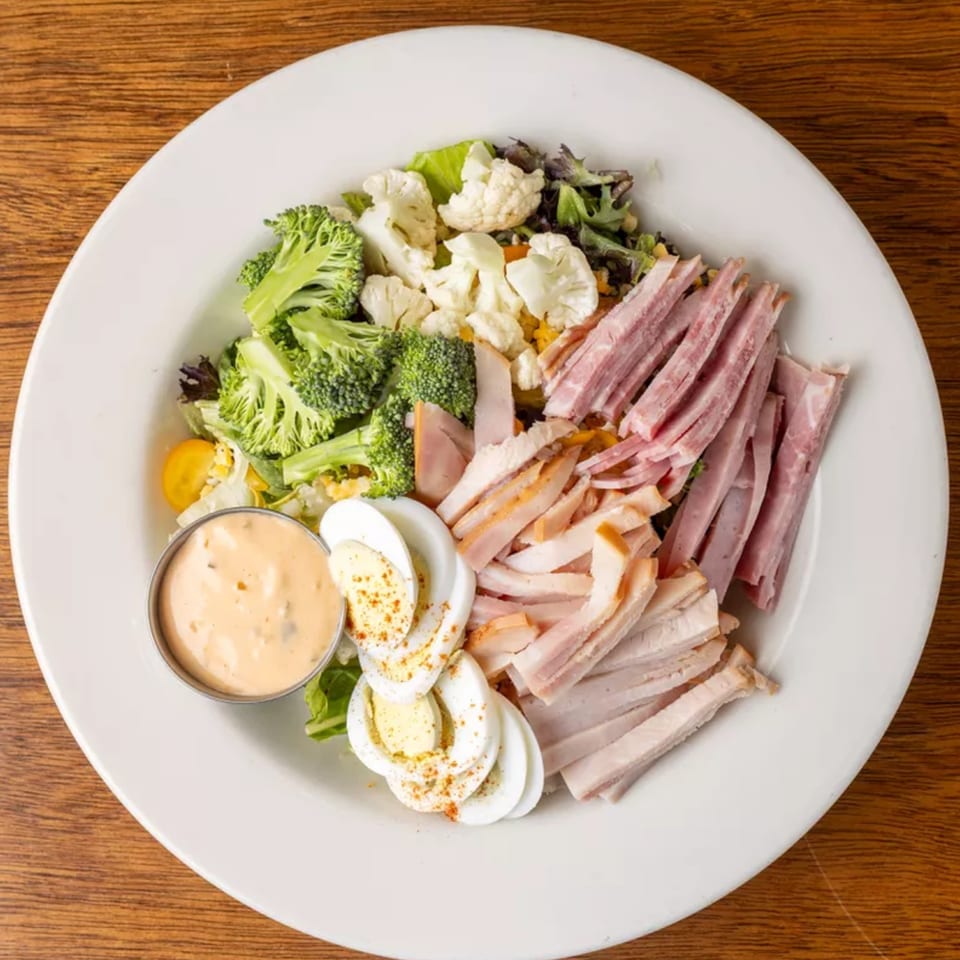 Overhead view of a chef salad with ham, turkey, broccoli, cauliflower, hard-boiled eggs, and dressing.