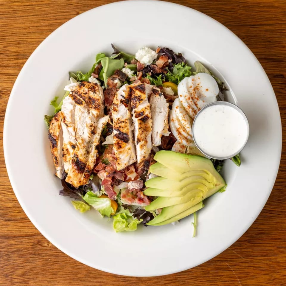 Grilled chicken salad with bacon, avocado, hard-boiled eggs, and a side of dressing.