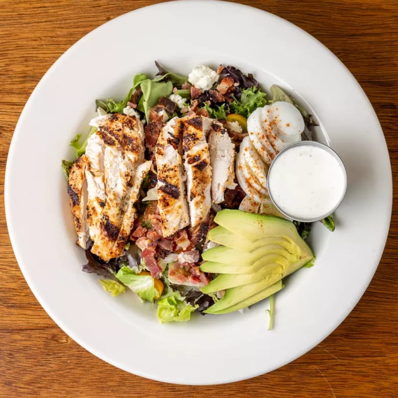 Grilled chicken salad with bacon, avocado, hard-boiled eggs, and a side of dressing.