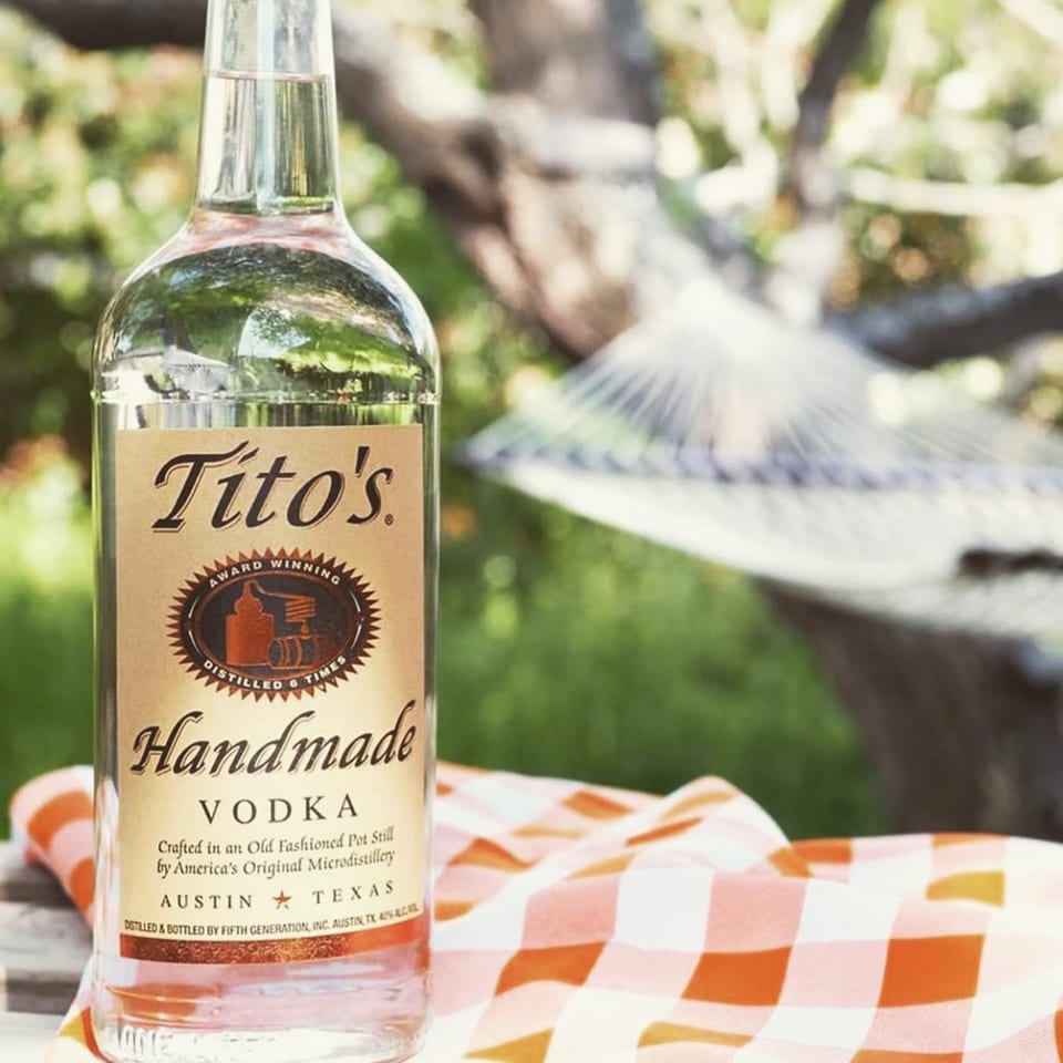 Tito's Handmade Vodka bottle on an orange and white checkered cloth outdoors with a hammock.